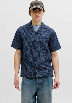 Mladý muž oblečený v modrej krátkorukáveovej košeli s vzorom, svetlomodrom tričku, sivej šiltovke s nápisom „Sunday Club“ a voľných modrých džínsoch na jednoduchom pozadí.