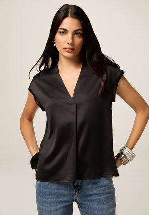 Zwarte satijnen blouse met een V-hals, kapmouwen en een ontspannen pasvorm, gecombineerd met blauwe jeans en gelaagde metalen armbanden om de pols.