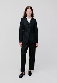 Zwart maatpak met een diepe V-hals, drie knopen en twee voorzakken. Gecombineerd met platte zwarte schoenen met een gladde textuur.