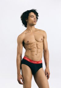 Fekete pamut férfi alsónadrág piros elasztikus derékkal, amelyet a "Calvin Klein" márkajelzés díszít. A modellen tónusos testalkat és göndör haj látható.