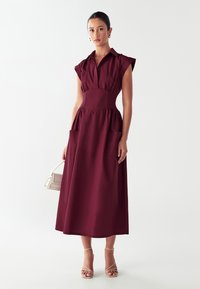 Abito bordeaux di lunghezza media con maniche corte, colletto, vita arricciata e due tasche frontali; tessuto liscio con design strutturato.