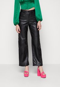 Černé kožené kalhoty širokého střihu s předními švy, v kombinaci s jasně růžovými vysokými sandály a zelenou saténovou blůzou s vycpanými rukávy.