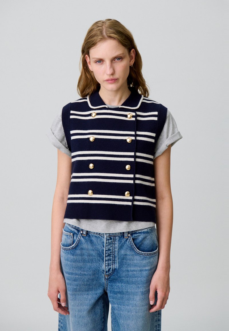 Gilet blu navy con strisce bianche, design doppiopetto, bottoni dorati e colletto, indossato sopra un T-shirt grigia, abbinato a jeans azzurri chiari.