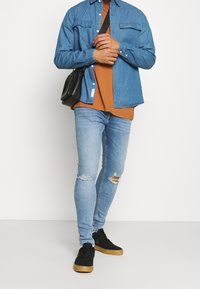 Camisa de mezclilla en azul claro, con diseño abotonado y dos bolsillos en el pecho. Lleva puesta sobre una camiseta naranja. Vaqueros azules ligeramente desgastados. Zapatillas negras.