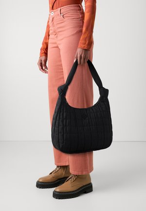 Sac à épaule noir matelassé avec une poignée douce, porté par une personne en pantalon en denim rose clair et bottes à lacets marron. Design simple et fonctionnel.