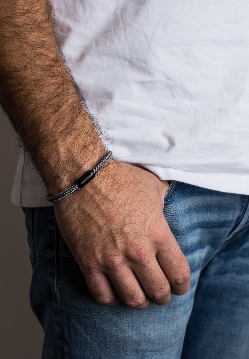 Sivý zapletený náramok s čiernym zapínaním, ktorý sa vyznačuje elegantným dizajnom. Nosený na zápästí na pozadí modrého denimového materiálu a bielej látky.