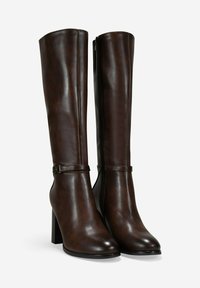 Botas de cuero marrón oscuro hasta la rodilla con tacones de bloque, punteras redondas, cremalleras laterales y correas decorativas alrededor de los tobillos sobre un fondo blanco.