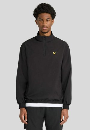 Giovane uomo che indossa un maglione nero con una cerniera a un quarto e un logo giallo a forma di uccello, in piedi davanti a uno sfondo grigio uniforme.