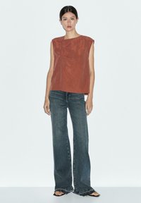 Rostfärgad ärmlös topp med asymmetrisk necklinje och knappring, paired med löst sittande mörk denimjeans och svarta sandaler.