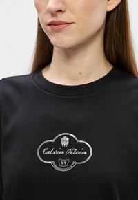 Zwarte katoenen t-shirt met een ronde halslijn en korte mouwen, met een zilver geborduurd logo met bloemaccenten en tekst.