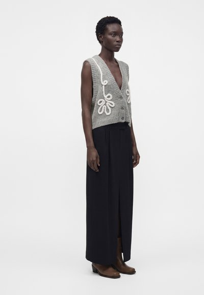 Femme debout portant un gilet sans manches gris en maille avec broderie blanche, une longue jupe noire fendue sur le devant et des bottines marron.