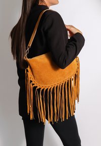 Suede schoudertas in mosterdgeel met franjes aan de onderkant, cirkelvormig snit en een enkele schouderband. Glad materiaal en minimale hardware.