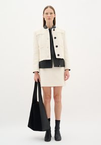 Giacca bianca a trama con colletto in pelle nera, abbinata a una mini gonna bianca. La modella tiene una borsa tote nera e indossa stivali alla caviglia neri.