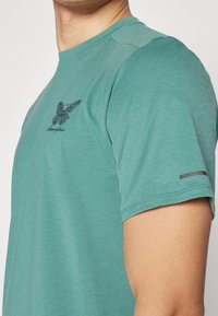 Syvänsininen lyhythihainen t-paita, jossa on teksturoitu kangas, pyöreä kaula-aukko ja musta graafinen printti edessä, sekä hopeinen yksityiskohta hihassa.