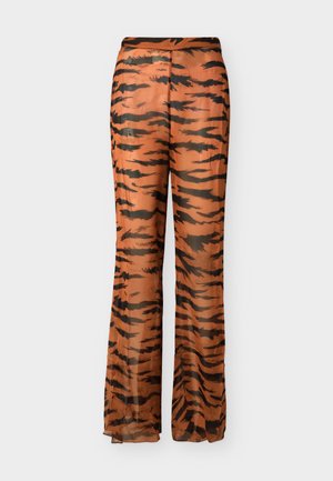 Mujer con top de bikini negro, pantalones anchos y transparentes con estampado de rayas de tigre, llevando un bolso grande de mimbre y calzando sandalias negras y gafas de sol.