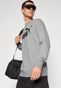 Grå sweatshirt med lange ærmer og et sort Puma-logo på forsiden, parret med en sort skuldertaske. Modellen bærer oversize sorte solbriller.