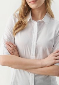 Bijela košulja s gumbima i kratkim rukavima, izrađena od lagane tkanine. Ima klasičnu kragnu i glatku, suptilnu teksturu.