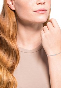 Pulsera de plata con una pequeña piedra preciosa azul, llevada en una muñeca de piel clara. El modelo tiene el cabello largo y ondulado de color rojo y viste una blusa beige.