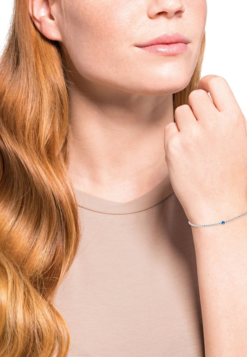 Pulsera de plata con una pequeña piedra preciosa azul, llevada en una muñeca de piel clara. El modelo tiene el cabello largo y ondulado de color rojo y viste una blusa beige.