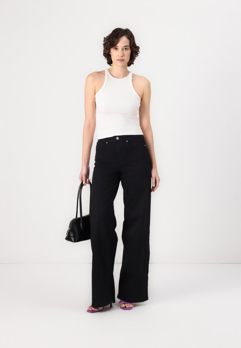 Jeans anchos de talle alto negros, mezcla de algodón suave, combinados con una camiseta sin mangas blanca de canalé. La modelo lleva un pequeño bolso negro y calza tacones púrpuras.