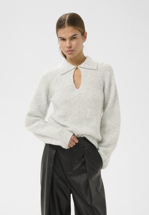 Giovane donna con capelli tirati indietro che indossa un maglione lavorato a maglia grigio chiaro e pantaloni in pelle neri a vita alta, che posa con un'espressione seria.