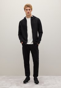 Felpa nera con zip e cordini, t-shirt bianca sotto, pantaloni jogger neri con tasca cargo e sneakers nere, su uno sfondo neutro.