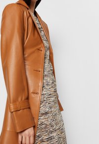 Cappotto in pelle marrone con un design aderente, chiusura a bottoni e maniche con cintura, sovrapposto a un vestito a fantasia con toni neutri e texture.