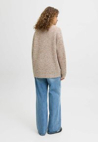 Gruby dziany kardigan w beżowym kolorze z kolorowymi plamkami, w oversize'owym kroju i prążkowanej strukturze. Połączony z jasnoniebieskimi spodniami typu wide-leg.