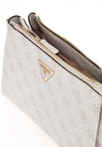 Beige Stoffbeutel mit strukturiertem Logomuster, zwei Reißverschlüssen und einem dreieckigen Logo-Akzent in Goldoptik. Glattes Innenfutter.