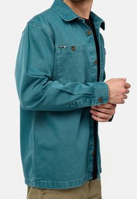 Giacca di jeans teal con chiusura a bottoni, due tasche sul petto e colletto classico. Presenta bottoni marroni e una vestibilità rilassata.