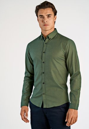 Mann trägt ein eng anliegendes, langärmliges grünes Hemd mit Knöpfen und dunkle Hose, steht vor einem schlichten hellen Hintergrund.