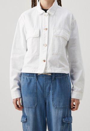 Person iført en hvid jakke med knapper og brystlommer samt løse blå denim cargobukser med for- og sidelommer.