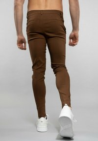 Pantalon slim marron en tissu lisse, avec un design de poche arrière. Associé à des baskets blanches, vu de l'arrière.