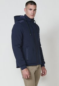 Felpa con zip di colore blu navy, realizzata in tessuto testurizzato, con cappuccio regolabile tramite cordino e tasche laterali. Presenta pannelli a contrasto e polsini a coste.