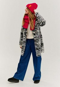 Šedý a čierny kabát so zebrovým vzorom na krémovom pletenom svetri, skombinovaný s širokými modrými denimovými nohavicami, červenou rebrovanou čiapkou a zodpovedajúcim šálom.