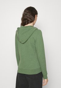 Žena s dlouhými vlnitými vlasy má na sobě zelenou mikinu s kapucí a černé džíny, stojí zády k jednolitému bílému pozadí.