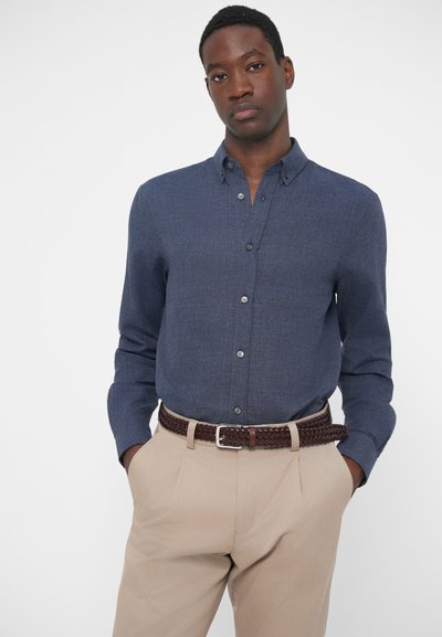 Camisa azul-marinho de botão com um acabamento texturizado, apresentando um colarinho clássico e um único bolso no peito, combinada com calças bege claras e um cinto trançado castanho.