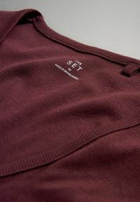 Bordeauxfarbener gerippter Stoff mit weicher Textur. Das Etikett trägt die Aufschrift "THE SET M MADE IN BANGLADESH" in Weiß und befindet sich am hinteren Kragen.