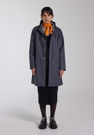 Grå regnjacka med huva, tryckknappar och dragsko-accenter. Bärs över en svart klänning, ihop med svarta klumpiga skor och en orange scarf.