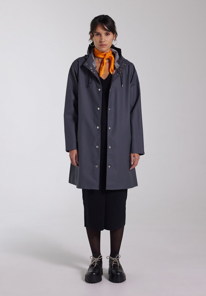 Grijze regenjas met een capuchon, drukknoopsluitingen en trekkoorden. Draagt over een zwarte jurk, gecombineerd met zwarte chunky schoenen en een oranje sjaal.