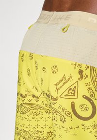 Pantalones cortos amarillos con un diseño impreso que presenta gráficos marrones. La cinturilla es beige con acentos texturizados y un gancho decorativo.