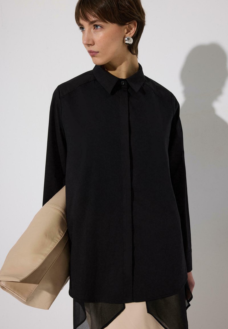 Femeie purtând o cămașă neagră cu mâneci lungi, nasturii închiși, cu guler, ținând o jachetă bej și purtând cercei mari argintii, uitându-se spre stânga.