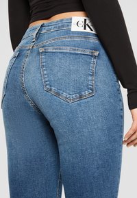 Blå denimjeans med hög midja, femfickarsdesign och synliga sömnadsdetaljer. Etikett visar "Calvin Klein Jeans"-logotypen.