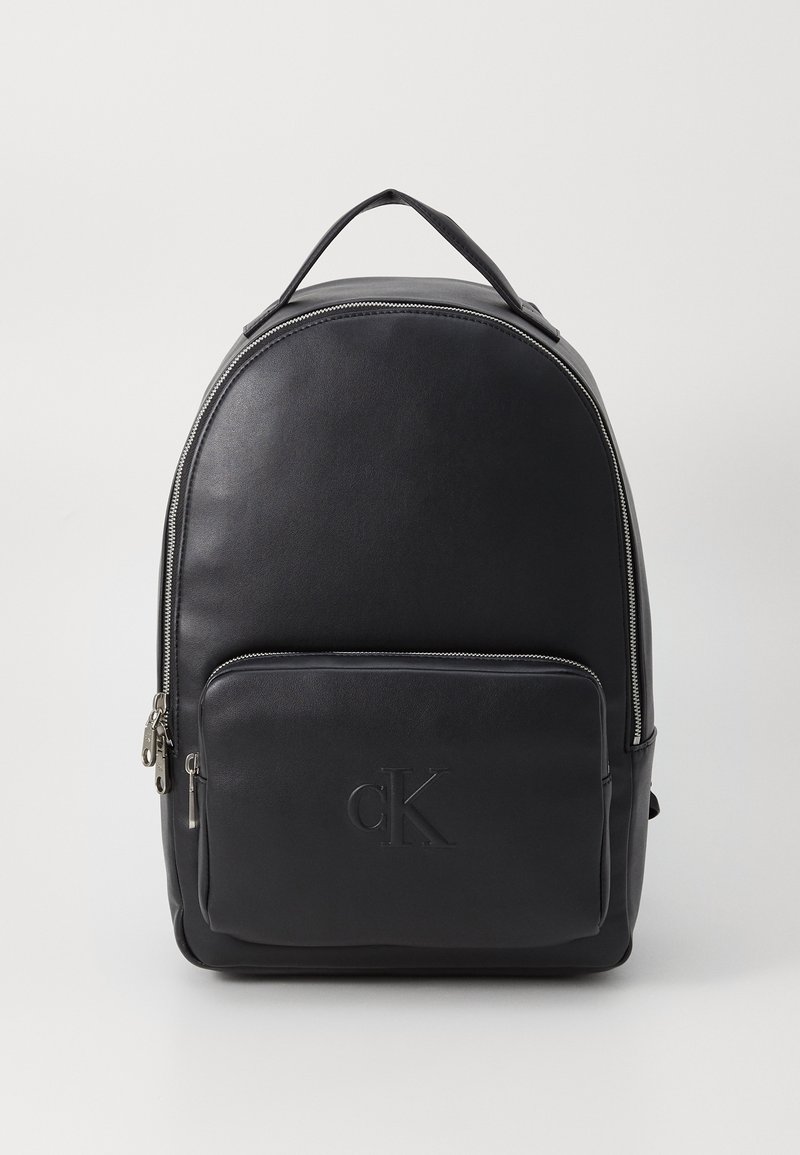 Svart läderryggsäck med rundad form, dragkedja i huvudfacket, frontficka och präglad logotyp. Har ett handtag på toppen och justerbara remmar.