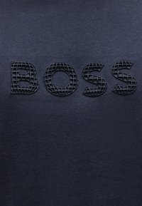 Μαύρα υφασμάτινα γράμματα που σχηματίζουν τη λέξη "BOSS" σε σκούρο γκρι φόντο, τα γράμματα έχουν σχέδιο πλέγματος και τρισδιάστατο εφέ.