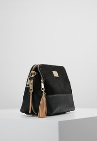 Czarna torba na ramię z zamszu i skóry ze złotymi elementami, podwójnym zamkiem i beżowym detalem z frędzlami. Prostokątny kształt o gładkiej fakturze.