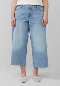 Jasnoniebieskie dżinsy typu wide-leg z surowym wykończeniem, pięć kieszeni i subtelne przetarcia. Połączone z białymi meshowymi butami slip-on.