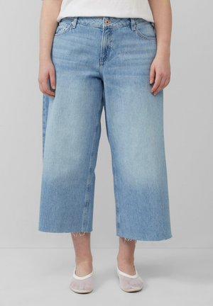 Helleblaue weit geschnittene Jeans mit rohem Saum, Fünf-Taschen-Design und subtiler Ausbleichung. Kombiniert mit weißen Netzschlupfschuhen.