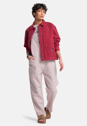 Rote Baumwolljacke mit Knopfverschluss und Vordertasche, kombiniert mit gestreiften Latzhosen in Weiß und Rot, getragen mit braunen Sandalen.