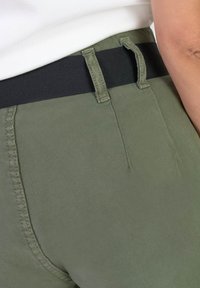 Pantalons vert olive avec une taille élastique noire, dotés de poches latérales et de détails de couture visibles sur une texture de tissu lisse.